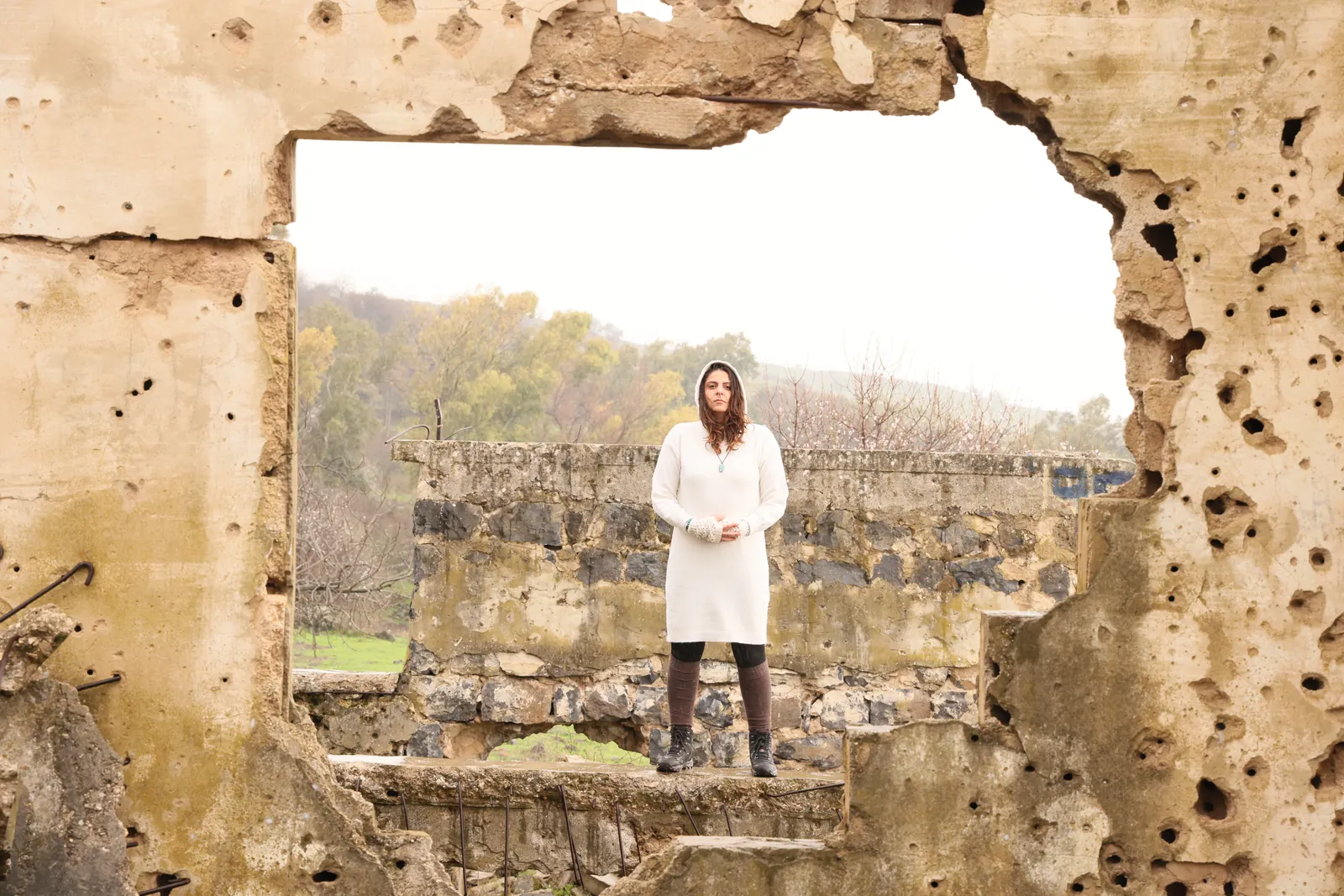 Rawan in white, seen through a broken, pitted wall of an abandoned village in the Golan Heights
